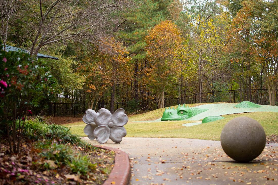 Climbable playground sculpture of a green dragon emerging from a hillside wtih a sidewalk sphere and bench in the foreground