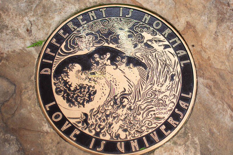 A round sidewalk paver with an intricate image of a wave crashing and swirling clouds overhead, with the text "Different is normal, love is universal' engraved around.
