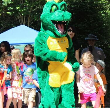 Walkaround character of Katal, the Kids Together Playground mascot who is a green and yellow dragon, leading a parade of children